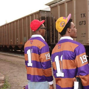 May include: Two people wearing matching purple and brown striped rugby shirts with the number "1" and "19" on the back. They are wearing baseball caps, one red and one yellow. The background includes a train and a cloudy sky.