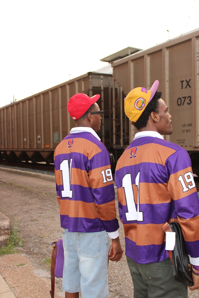 May include: Two people wearing matching purple and brown striped rugby shirts with the number "1" and "19" on the back. They are wearing baseball caps, one red and one yellow. The background includes a train and a cloudy sky.