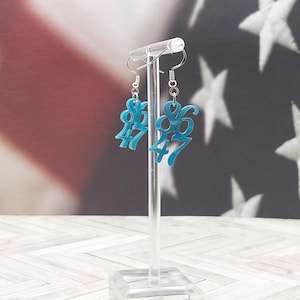 May include: A pair of turquoise earrings with the numbers "86" and "47" in a stylized font. The earrings hang from silver-colored hooks. The background features a blurred American flag.