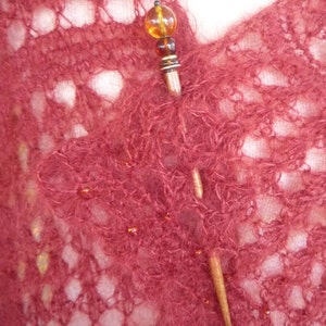 May include: A close-up of a burgundy knitted shawl with a wooden shawl pin. The pin has a round amber bead at the top and a smaller amber bead below it.