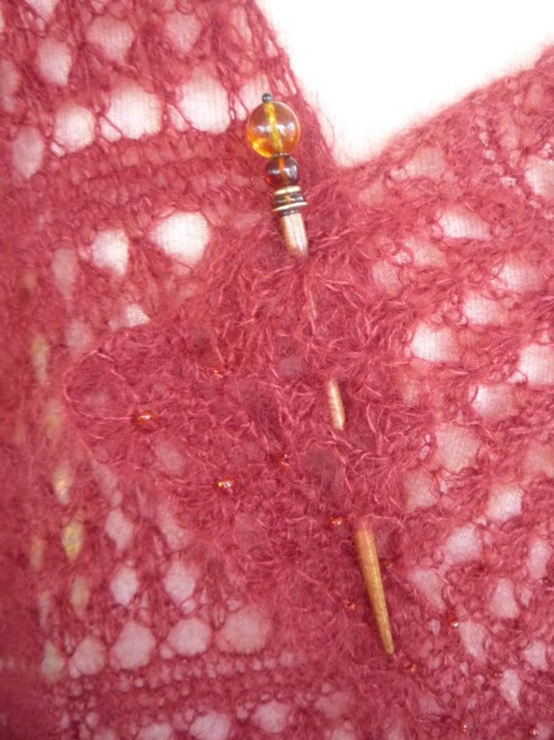 May include: A close-up of a burgundy knitted shawl with a wooden shawl pin. The pin has a round amber bead at the top and a smaller amber bead below it.