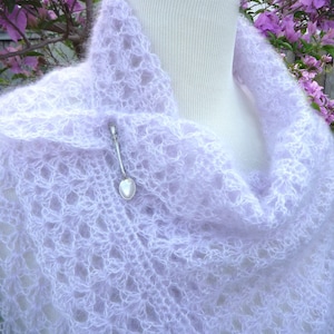 Peut inclure: Un châle en crochet délicat de couleur mauve clair avec un motif en dentelle. Le châle est drapé sur un mannequin blanc et a un pendentif en forme de cœur argenté attaché.