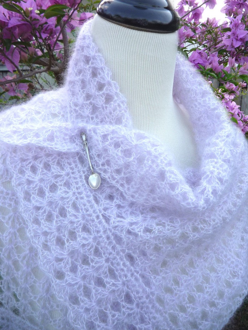 May include: A delicate, light purple crochet shawl with a lacy pattern. The shawl is draped over a white mannequin and has a silver heart-shaped pendant attached.