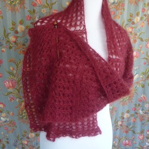 May include: A burgundy lace shawl with a delicate openwork pattern. The shawl is draped over a white dress form and is made of a soft, lightweight yarn.