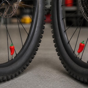 Puede incluir: Dos tapones de válvula con forma de chile rojo fijados a los neumáticos de goma negra de una bicicleta. Cada tapón presenta un cuerpo rojo brillante con un pequeño tallo verde en la parte superior, asegurado sobre las válvulas metálicas.