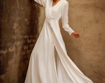 May include: A white, long-sleeved dress with a V-neckline and a high slit. The dress features a button-down front and a flowing skirt. The fabric appears to be a soft, lightweight material, suitable for formal occasions.