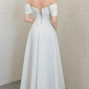 May include: A white off-the-shoulder wedding dress with a lace-up back. The dress has short sleeves and a flowing skirt. The model is standing with their back to the camera.