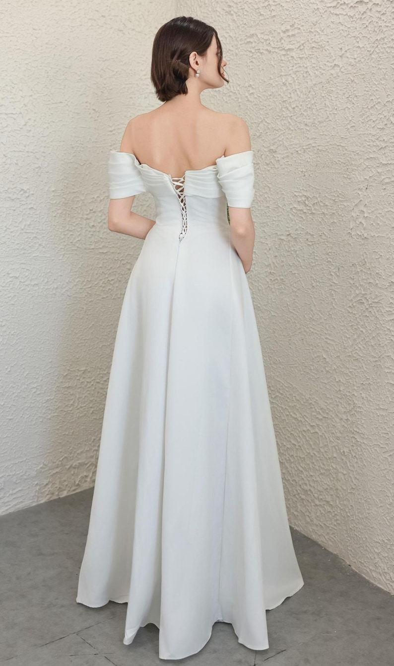 May include: A white off-the-shoulder wedding dress with a lace-up back. The dress has short sleeves and a flowing skirt. The model is standing with their back to the camera.