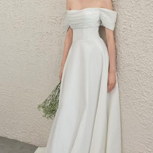 May include: A white, off-the-shoulder wedding dress with a flowing A-line skirt. The bodice has delicate pleating, and the bride holds a small bouquet of greenery. The dress is made of a smooth, elegant fabric.