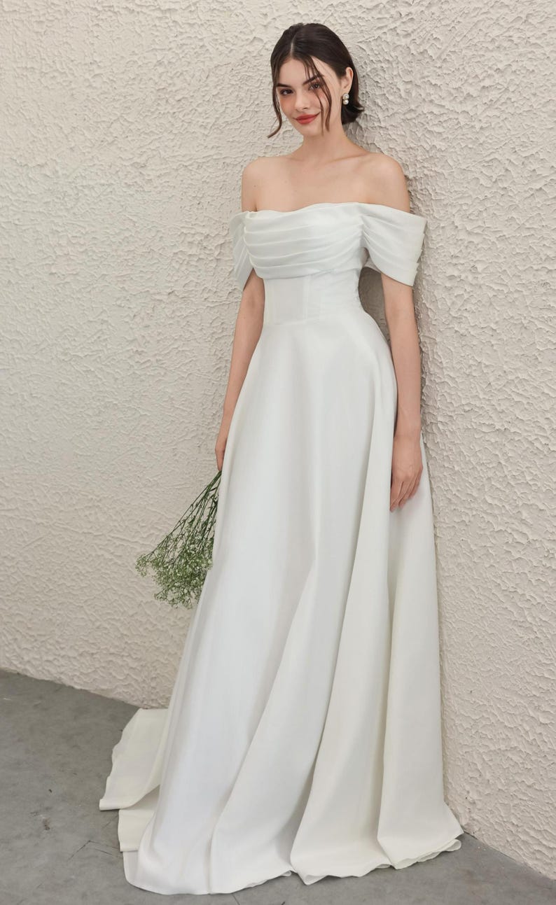May include: A white, off-the-shoulder wedding dress with a flowing A-line skirt. The bodice has delicate pleating, and the bride holds a small bouquet of greenery. The dress is made of a smooth, elegant fabric.
