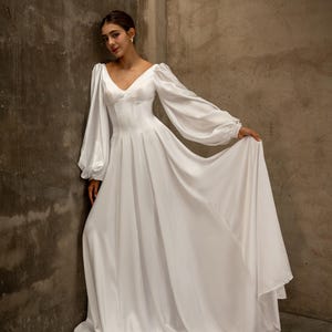 Op de afbeelding: Een witte satijnen jurk met een V-hals en lange, volumineuze mouwen. De jurk heeft een getailleerd lijfje en een zwierige rok. Het model leunt tegen een getextureerde muur, wat het elegante ontwerp van de jurk benadrukt. Geschikt voor bruiloften en formele evenementen.