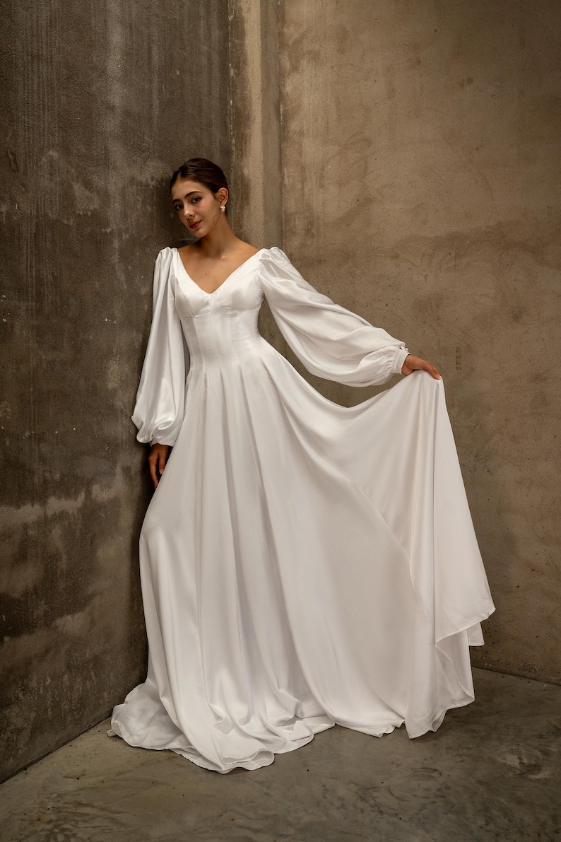 Op de afbeelding: Een witte satijnen jurk met een V-hals en lange, volumineuze mouwen. De jurk heeft een getailleerd lijfje en een zwierige rok. Het model leunt tegen een getextureerde muur, wat het elegante ontwerp van de jurk benadrukt. Geschikt voor bruiloften en formele evenementen.