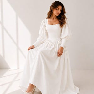 A woman in a long white dress with puffy sleeves and a high neckline stands in a studio setting, looking down at her hands.