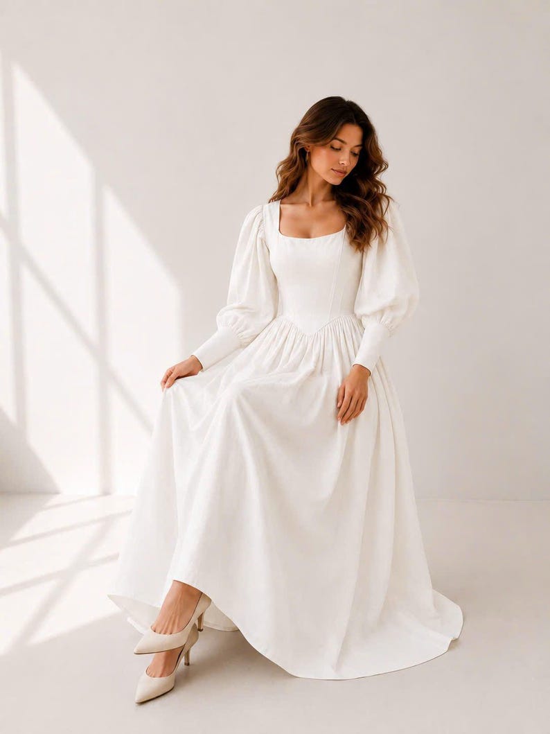 A woman in a long white dress with puffy sleeves and a high neckline stands in a studio setting, looking down at her hands.