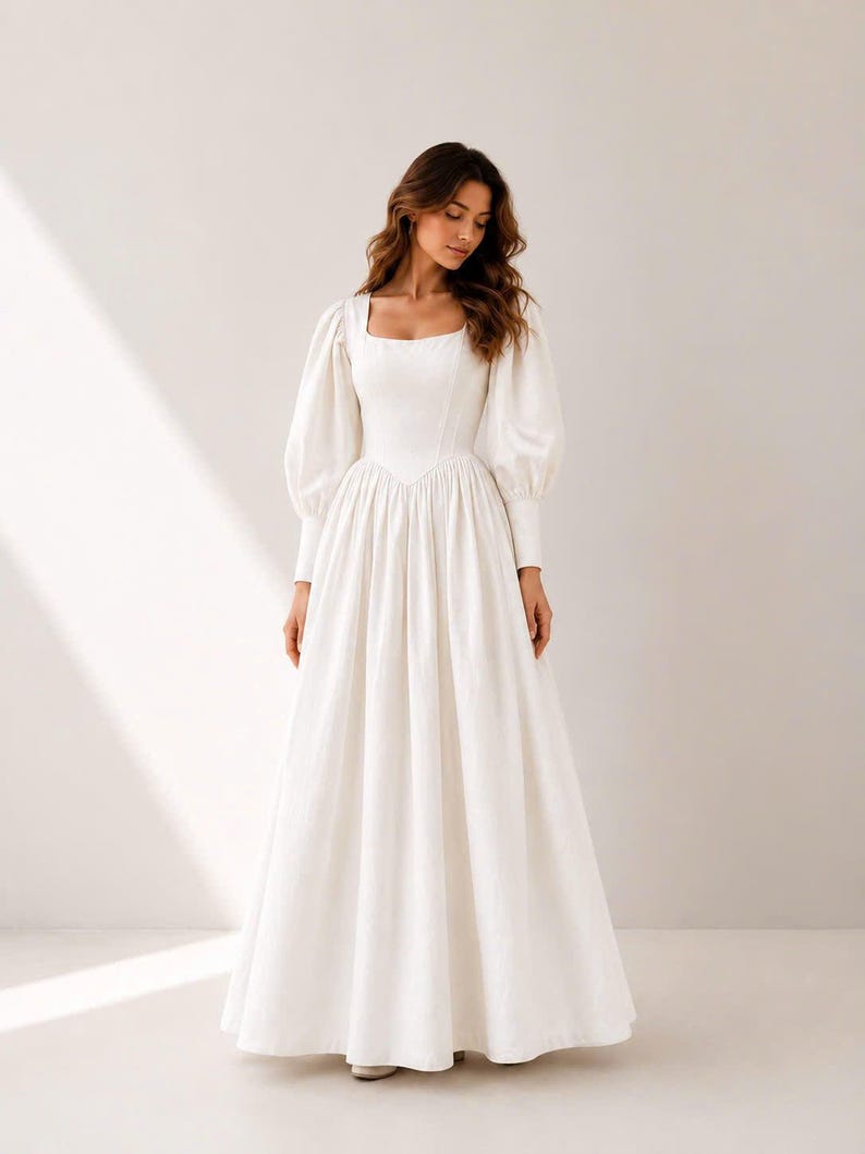 A woman wearing a long white dress with puffed sleeves stands in a studio setting.