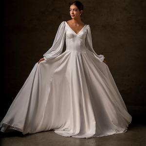 Op de afbeelding: Een witte satijnen trouwjurk met een V-hals en lange, vloeiende mouwen. De jurk heeft een getailleerd lijfje en een volle rok die vanaf de taille uitloopt. Het model draagt de jurk in een studio.