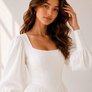 A young woman with long, wavy brown hair wearing a white dress with puffy sleeves, posing with her hand on her head.