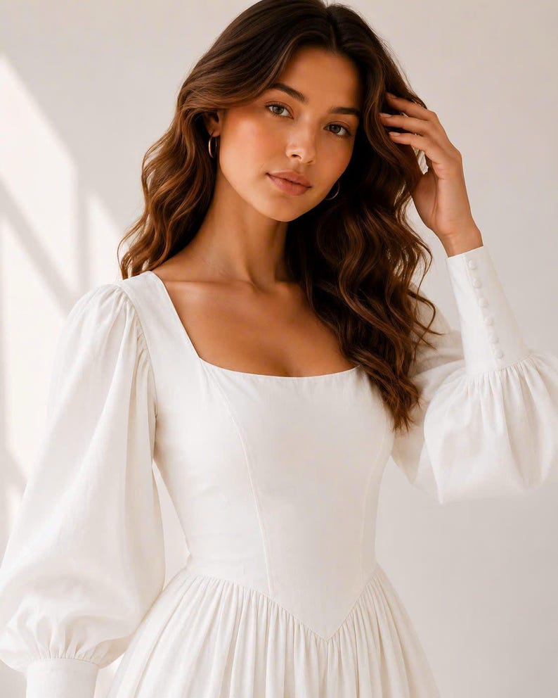 A young woman with long, wavy brown hair wearing a white dress with puffy sleeves, posing with her hand on her head.