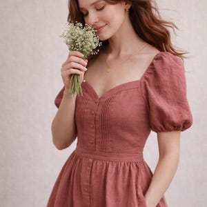 May include: A dusty rose-colored dress featuring a square neckline, short puff sleeves, and a button-down front bodice with vertical pleat detailing. The garment includes a fitted waistline and a gathered skirt with side pockets.