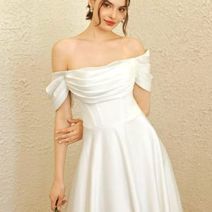 May include: A white off-the-shoulder wedding dress with a fitted bodice and a flowing skirt. The dress has a draped neckline and short sleeves. The bride is holding a small bouquet of greenery.