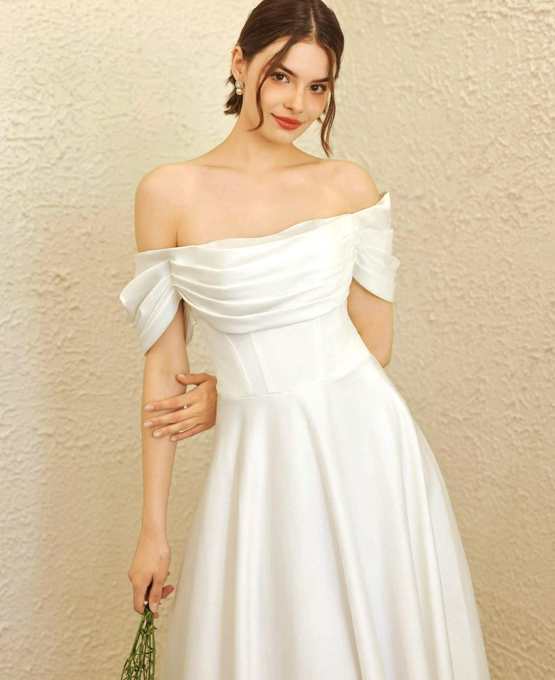 May include: A white off-the-shoulder wedding dress with a fitted bodice and a flowing skirt. The dress has a draped neckline and short sleeves. The bride is holding a small bouquet of greenery.