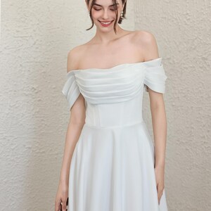 May include: A white, off-the-shoulder wedding dress with a fitted bodice and flowing skirt. The dress has a pleated design on the bodice and short sleeves. The bride is holding a small bouquet of greenery.