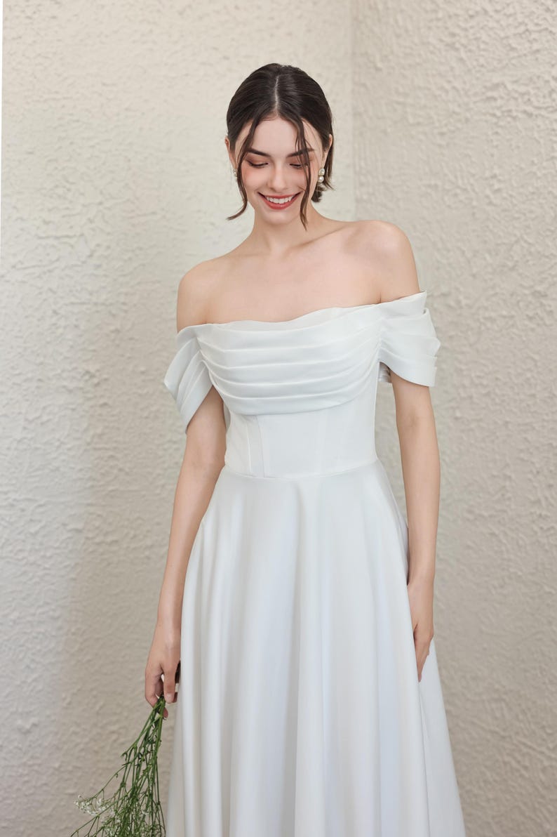 May include: A white, off-the-shoulder wedding dress with a fitted bodice and flowing skirt. The dress has a pleated design on the bodice and short sleeves. The bride is holding a small bouquet of greenery.