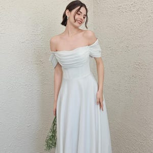 May include: A white off-the-shoulder wedding dress with a fitted bodice and flowing skirt. The dress features short sleeves and is held by a person. The person is holding a small bouquet of greenery.
