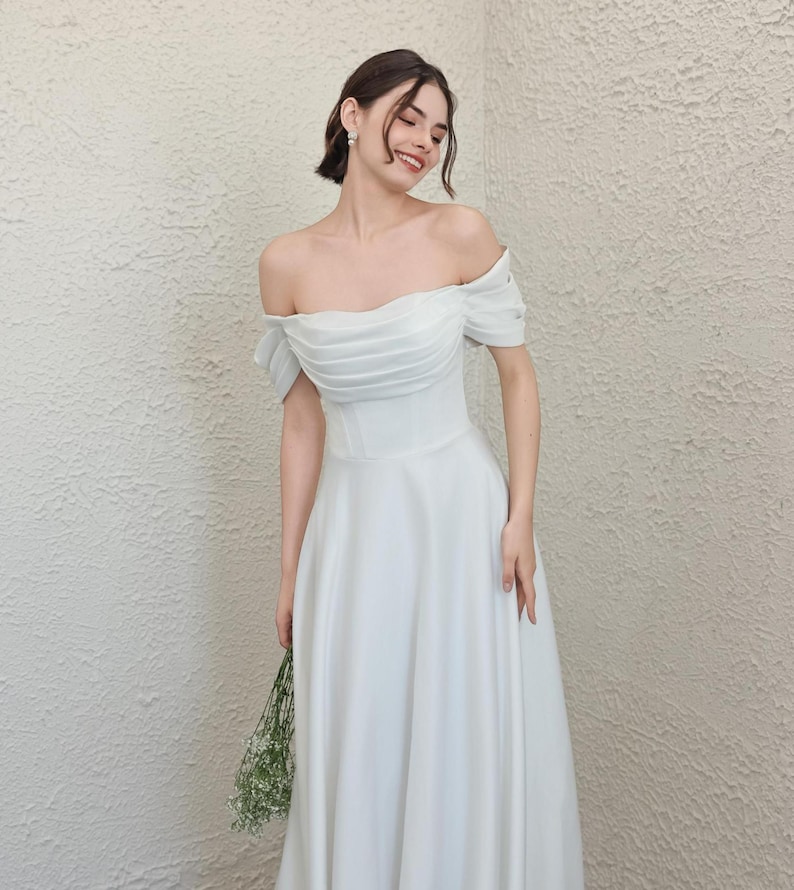 May include: A white off-the-shoulder wedding dress with a fitted bodice and flowing skirt. The dress features short sleeves and is held by a person. The person is holding a small bouquet of greenery.