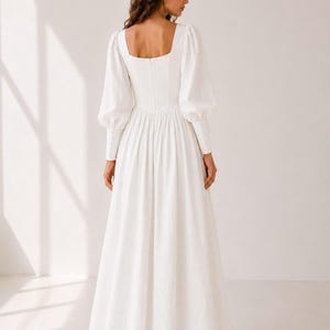 A woman wearing a long white dress with puffy sleeves stands facing away from the camera.