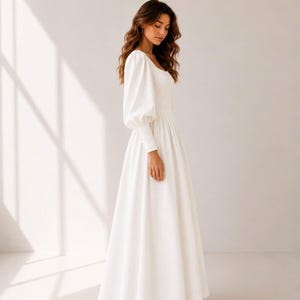 A woman in a long white dress stands in a studio, her gaze directed downwards.
