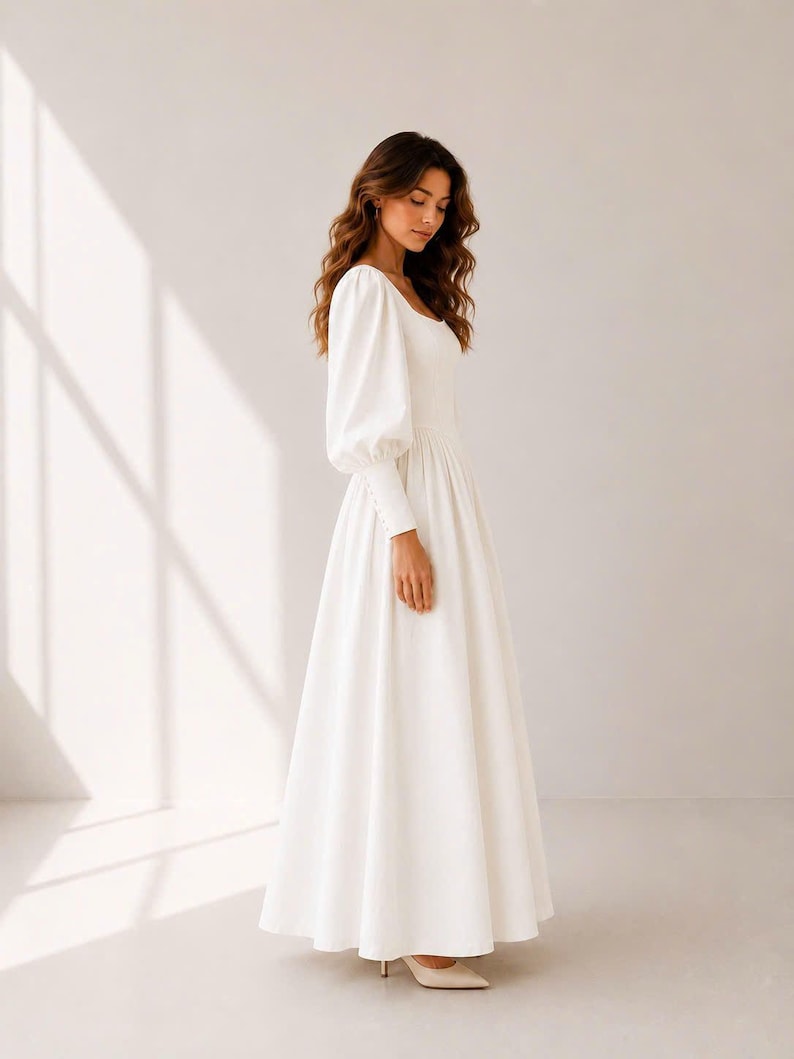 A woman in a long white dress stands in a studio, her gaze directed downwards.