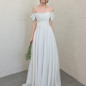 May include: A white off-the-shoulder wedding dress with a fitted bodice and a flowing A-line skirt. The dress features a ruched bodice and short sleeves. The person is holding a small bouquet of white flowers.