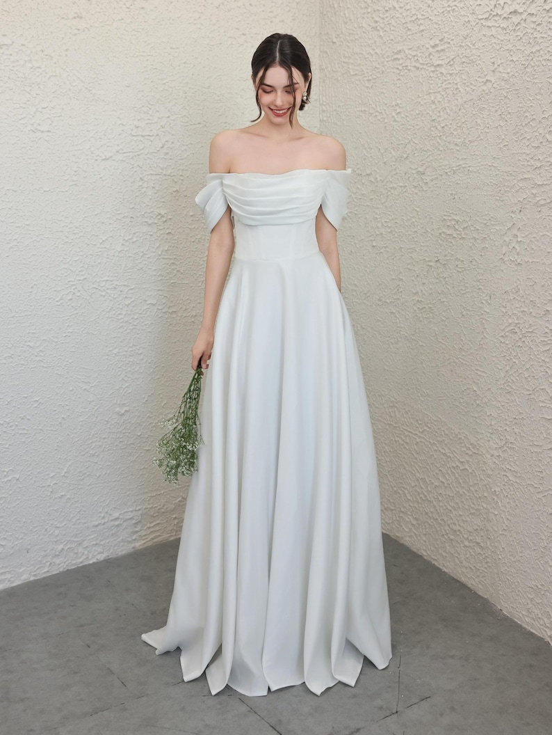 May include: A white off-the-shoulder wedding dress with a fitted bodice and a flowing A-line skirt. The dress features a ruched bodice and short sleeves. The person is holding a small bouquet of white flowers.