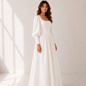 A woman wearing a long white dress with puffed sleeves stands in a studio setting.