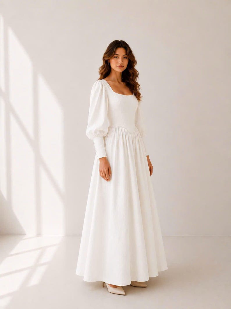 A woman wearing a long white dress with puffed sleeves stands in a studio setting.