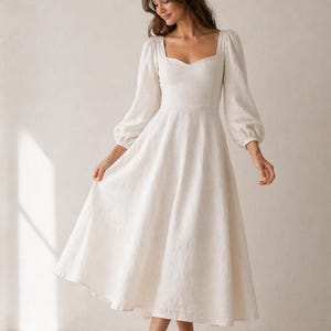 May include: A cream-colored midi dress featuring a sweetheart neckline, long puff sleeves with fitted cuffs, and a full, flowing skirt. The fabric has a subtle, textured pattern throughout, and the bodice is fitted to the waist.