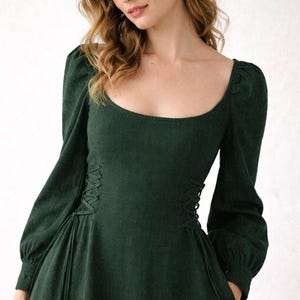 May include: A dark forest green long-sleeved dress featuring a square neckline, gathered puff sleeves with buttoned cuffs, and decorative lace-up corset-style detailing on both sides of the waist with matching green cord ties.