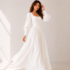 A woman wearing a long white dress with puffy sleeves and a high neckline stands in a studio setting, with her hand on her head.