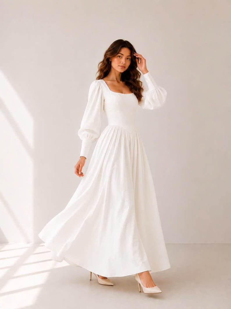 A woman wearing a long white dress with puffy sleeves and a high neckline stands in a studio setting, with her hand on her head.