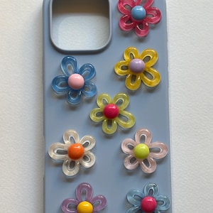 May include: A light blue phone case adorned with colorful, translucent flower embellishments. The flowers feature various colors, including pink, yellow, blue, and orange, with contrasting center beads. The case is designed to protect a phone.