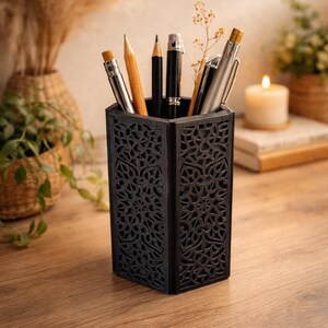 May include: A black, hexagonal pen holder with a decorative, openwork design. The holder contains various pens and pencils. The pen holder is made of a dark material and sits on a wooden surface. The background includes plants and a candle.