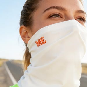 Puede incluir: Un cubrecuello blanco con la palabra "ME" bordada en naranja. El cubrecuello lo lleva una persona que lleva una camiseta sin mangas verde neón, con una carretera al fondo.