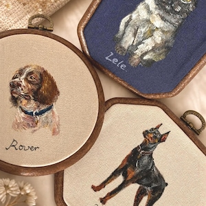 May include: Three custom pet portraits in wooden embroidery hoops. One round hoop features a brown dog named Rover. Two hexagonal hoops feature a grey cat named Zele and a black and tan dog named Kane. Each portrait is hand-painted on fabric.