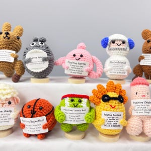 May include: A collection of handmade crochet figures, each with a unique design and holding a sign with positive affirmations. The figures include a kangaroo, Totoro, a kettle, an astronaut, a sheep, a basketball, a tennis ball, sunshine, and a chick.