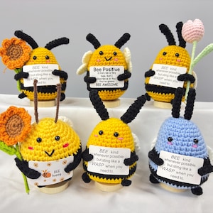 May include: A selection of handmade crochet bees in yellow and blue, with black stripes. Each bee holds a sign with positive messages and some hold flowers. The bees are displayed on a white surface.