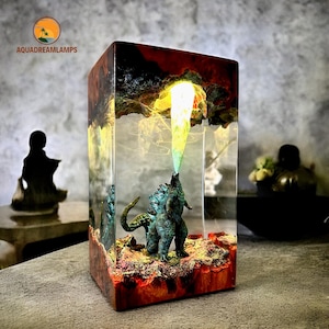 May include: A rectangular resin lamp featuring a Godzilla figurine emitting a yellow beam. The lamp has a wooden base and a fiery, cloud-like design inside. The text "AQUADREAMLAMPS" is visible on the top left.