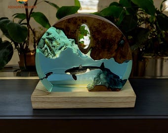 May include: A decorative lamp featuring a black and white orca swimming in a blue resin ocean scene. The lamp has a wooden base and a wooden frame with a natural wood grain. The lamp is illuminated from within, creating a warm glow.