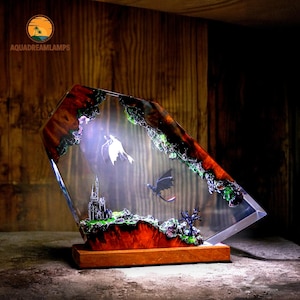 May include: A decorative resin lamp featuring a fantasy scene with dragons, a castle, and a tree. The lamp has a wooden base and a clear, angled resin body with a colorful interior. The lamp has the text "AQUADREAMLAMPS".