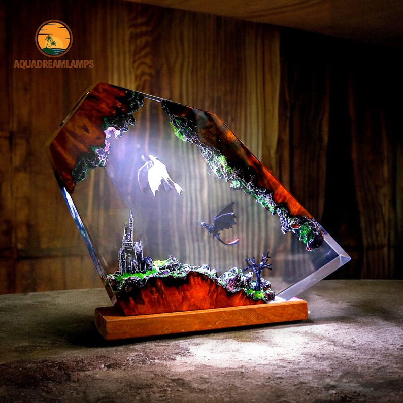 May include: A decorative lamp featuring a fantasy scene with two dragons, a castle, and a landscape encased in clear resin. The lamp has a wooden base and a unique, artistic design. The resin is accented with red, green, and brown colors.